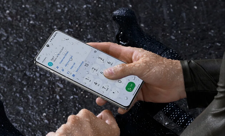 Ruce drží mokrý telefon OnePlus 13 5G, jehož displej s kapkami vody stále reaguje na dotyk a zobrazuje číselník hovoru.