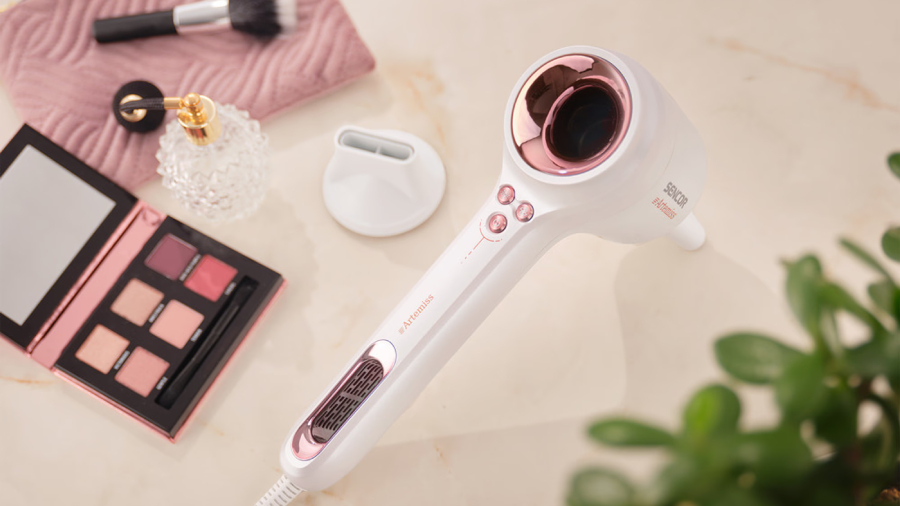 Vysoušeč vlasů Sencor SHD 9100RS Artemiss položený na stole, vedle něj je položený také difuzér a kolem toho, paletka stínů na oči, štěteček a parfém.