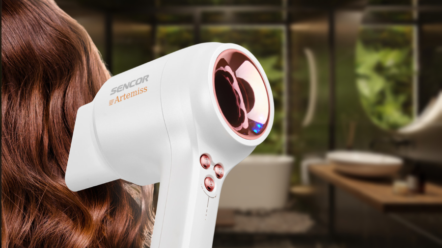 Detailní pohled na fén Sencor SHD 9100RS Artemiss, kde je vidět spíše jeho zadní část, kde se nachází LED diody a tlačítka. Fén má nasazený koncentrátor a míří na tmavě hnědé vlasy ženy.