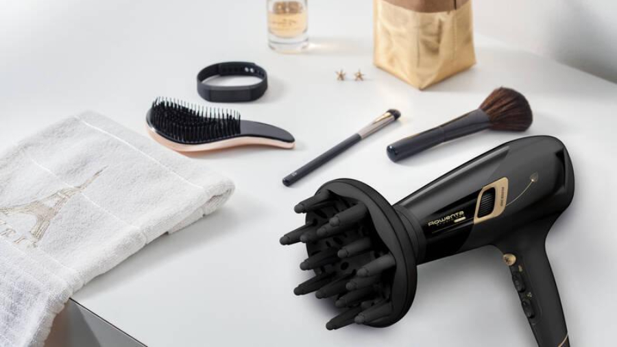 Rowenta Studio Dry Glow CV5831F0 v černém provedení s nasazeným difuzérem ležící na stole s ručníkem, kartáčem na vlasy a štětcemi na líčení.