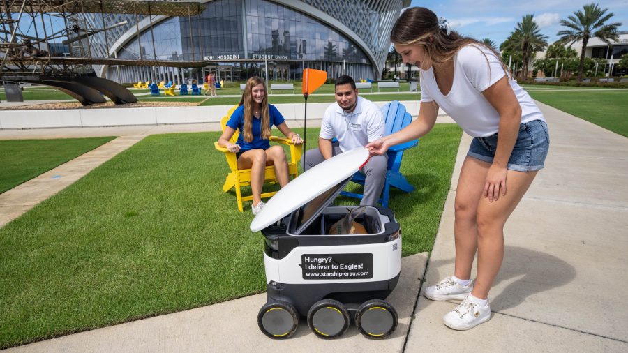 Robot Foodora od Starship doručuje jídlo studentce na univerzitním kampusu.