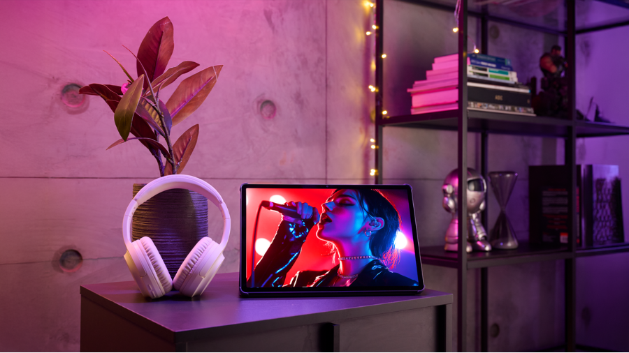 Tablet Lenovo Idea Tab 11" zobrazuje záběr zpěvačky s mikrofonem, postavený na stolku vedle bílých sluchátek a pokojové rostliny, v místnosti osvětlené růžovým a fialovým neonovým světlem.