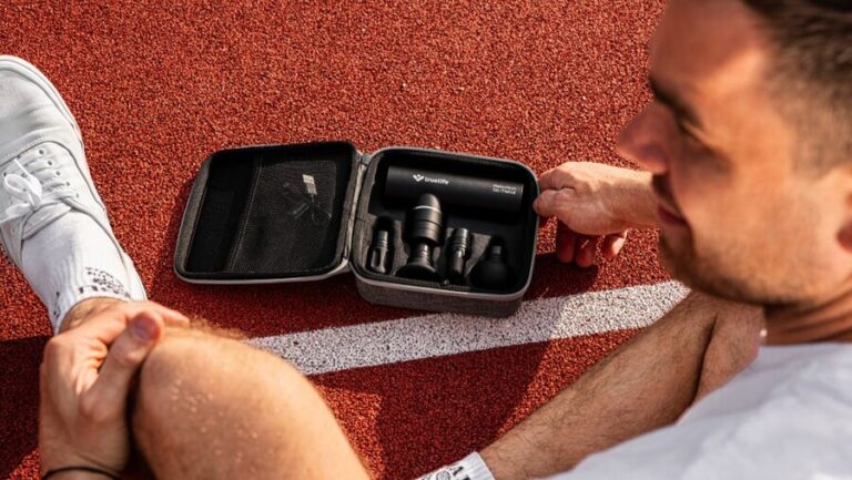 Muž sedící na atletické dráze si prohlíží přenosné pouzdro s černou masážní pistolí TrueLife RelaxGun G6 a sadou nástavců.