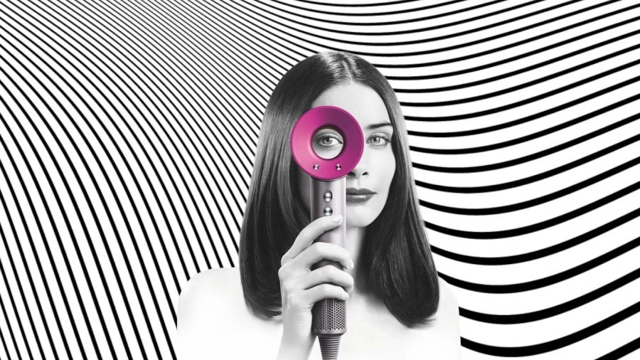 Černobílý obrázek ženy, která drží před sebou fén Dyson Supersonic HD07 v šedo-růžovém provedení a jedním okem kouká skrz jeho tunel v hlavici.