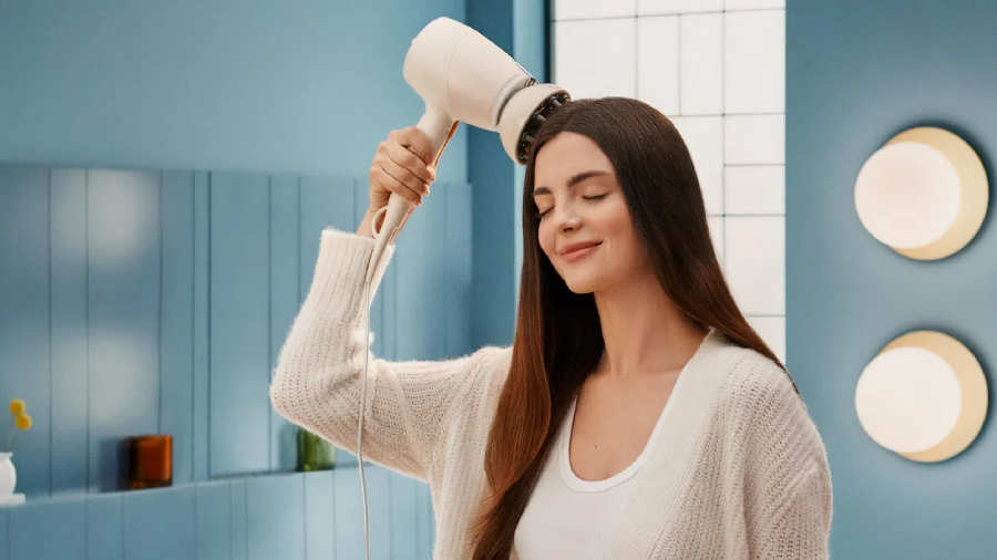 Žena s dlouhými hnědými vlasy v modré koupelně masírující si temeno hlavy fénem Philips Series 9000 SenseIQ, který má nasazený masážní nástavec.