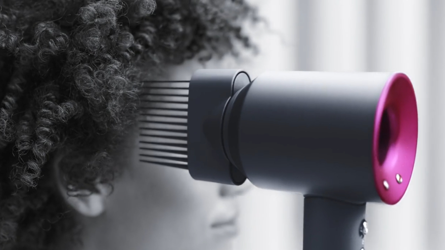 Fén Dyson Supersonic HD07 s nasazeným hřebenem se širokými zuby těsně u vlasů kudrnaté ženy.