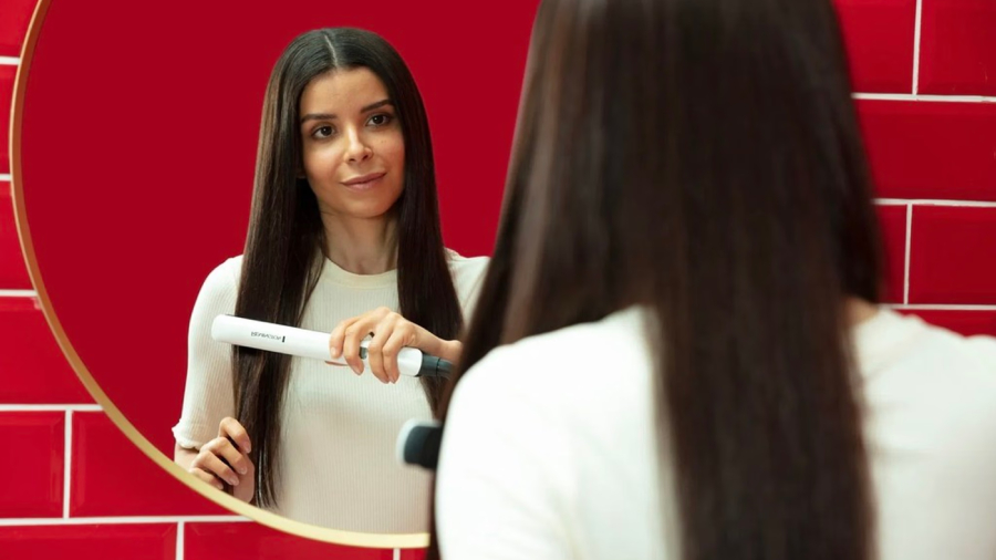 Žena s dlouhými hnědými vlasy stojící v červeně obložené koupelně před zrcadlem a žehlí si vlasy žehličkou Remington S8500 Shine Therapy.