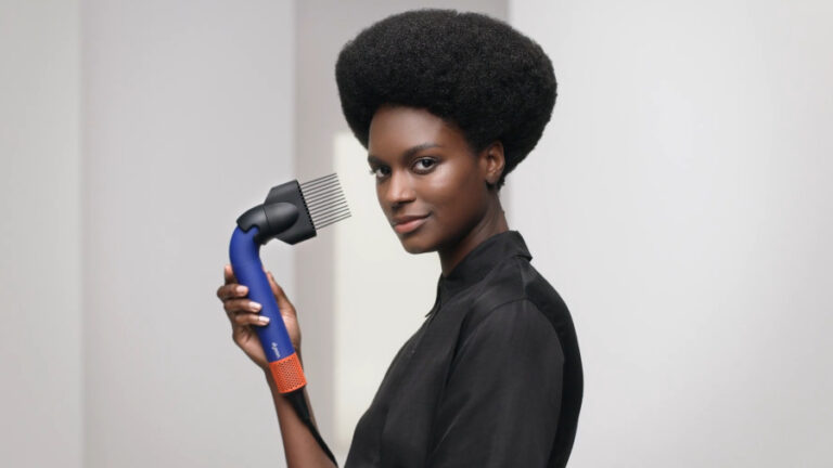 Černošská žena s hustými kudrnatými vlasy typu afro, která drží v ruce fén Dyson Supersonic r HD18 s nasazeným hřebenem se širokými zuby.