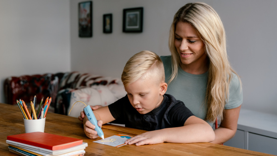 Chlapec v černém tričku kreslí modrým 3D perem LAMAX ArtKid1 pod dohledem dospělé ženy.