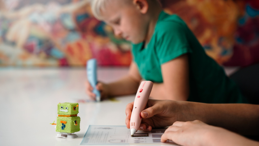 Dětské ruce používají růžové 3D pero LAMAX ArtKid1 k obkreslování šablony, vedle stojí hotový zelený 3D robot.