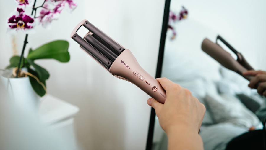 Ruka držící kulmu na vlasy TrueLife HairWaver W6, v pozadí jsou domácí rostliny s fialovo-bílými květy.