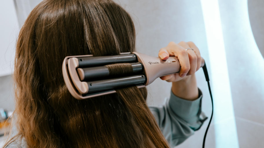 Detail hlavy ženy s dlouhými hnědými vlasy, která si vytváří vlny pomocí trojkulmy TrueLife HairWaver W6.