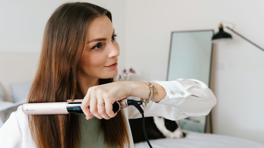 Žena s hnědými vlasy upravující si v pokoji vlasy žehličkou TrueLife HairIron N3.