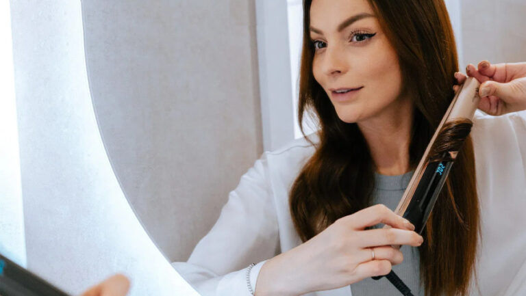 Žena s hnědými vlasy si před zrcadlem natáčí pramen vlasů žehličkou na vlasy TrueLife HairIron N3.