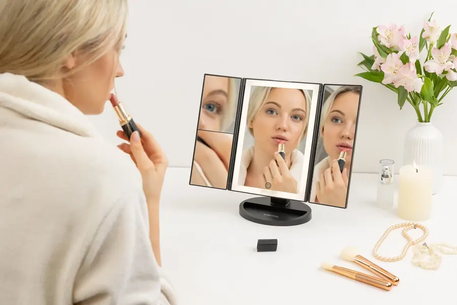 Tipy na vánoční dárky pro ženy: Žena v županu si nanáší rtěnku a dívá se do černého, trojdílného LED zrcátka Siguro LM-L751B Pure Beauty.