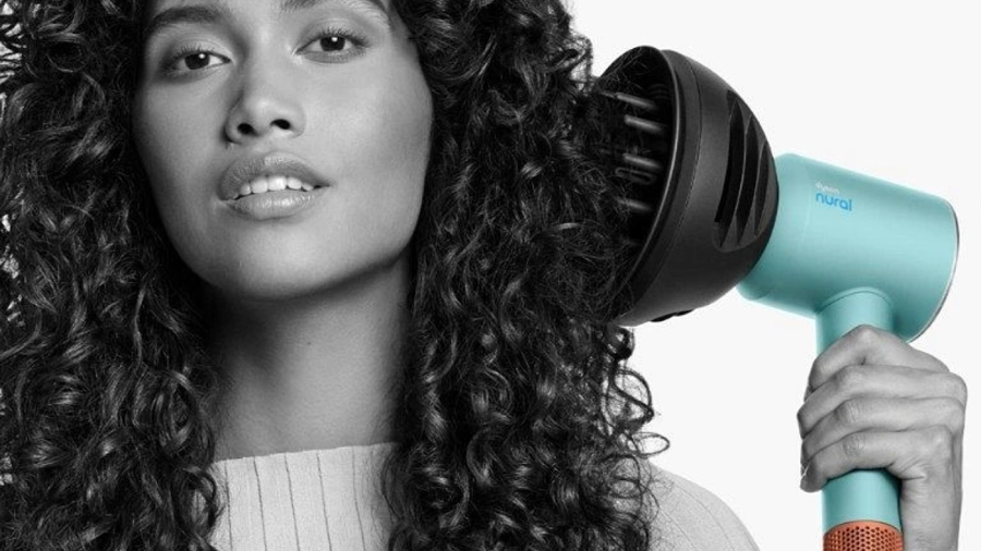 Žena s kudrnatými vlasy vyobrazená černobíle upravující si vlasy fénem Dyson Supersonic Nural s nasazeným difuzérem. Fén je vyobrazen v barevné kombinaci Curly + Coily Ceramic patina/Topaz orange, aby na obrázku vynikl.