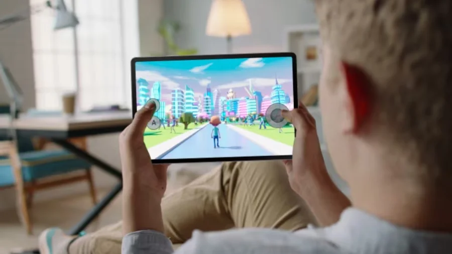 Mladý muž sedí a drží tablet Samsung Galaxy Tab S10 FE v horizontální poloze a hraje hru s barevnou 3D animovanou krajinou města.