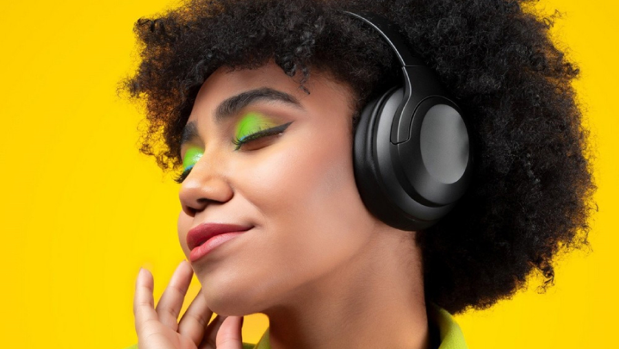 Žena se zavřenýma očima, výrazně nalíčenýma zeleno-žlutě, s nasazenými bezdrátovými sluchátky přes hlavu od značky Niceboy.