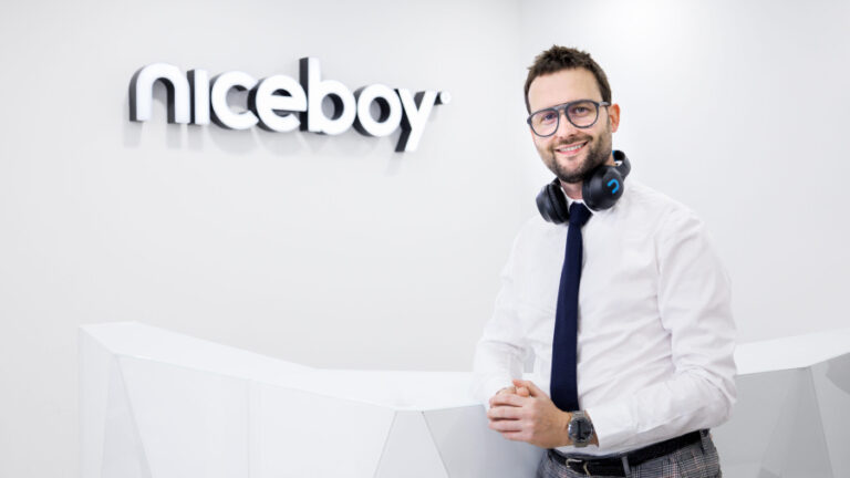 Ředitel značky Niceboy Michal Černý opřený o bílý pult v bílé košili s kravatou a sluchátky Niceboy zavěšenými kolem krku. Na zdi v pozadí je taktéž název značky Niceboy.