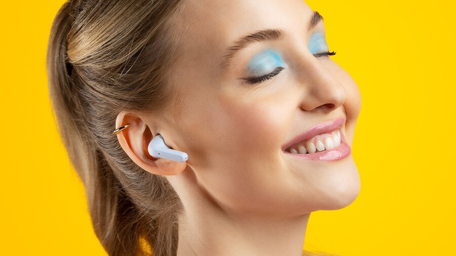 Tipy na vánoční dárky pro ženy: Detailní záběr na usměvavou ženu s modrými očními stíny, která má v uchu bílé bezdrátové sluchátko Niceboy HIVE Pins 3 ANC.