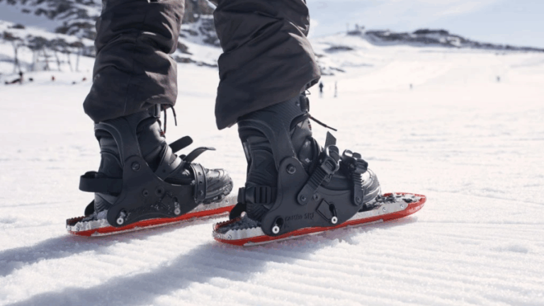 Vizionáři 2025: Detailní pohled na nohy osoby, která stojí na sněhu s nasazenými minilyžemi/sněžnicemi Comboski. Lyže jsou černé s červeným spodním okrajem, mají robustní vázání a kovové hroty. Osoba má na sobě tmavé lyžařské kalhoty.
