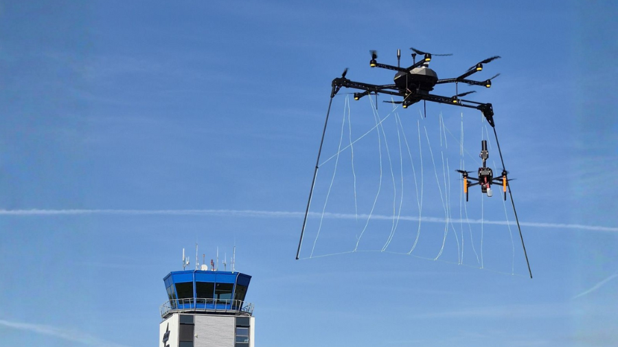 Vizionáři 2025: EAGLE.ONE "lovec dronů" svými lany chytá menší dron. V pozadí je vidět modrobílá letištní řídící věž a modrá obloha s kondenzačními čárami.