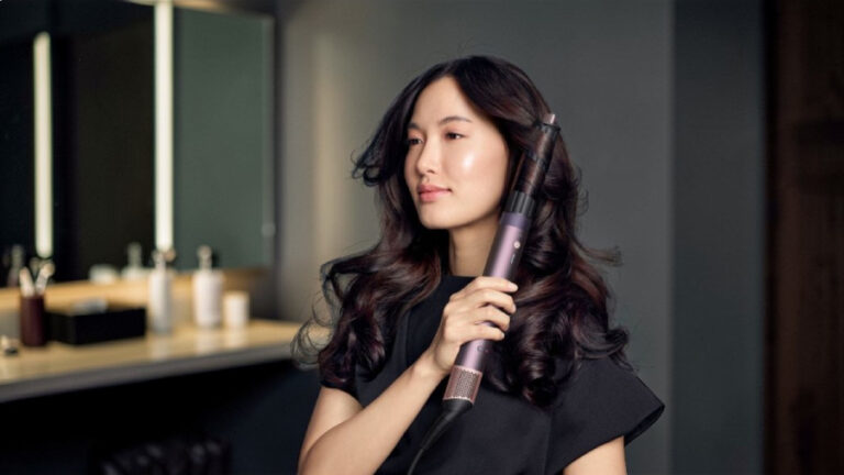 Mladá žena s tmavými vlnitými vlasy používá kulmofén Dyson Airwrap Co-anda 2x™ k vytvoření objemných loken v moderní koupelně.