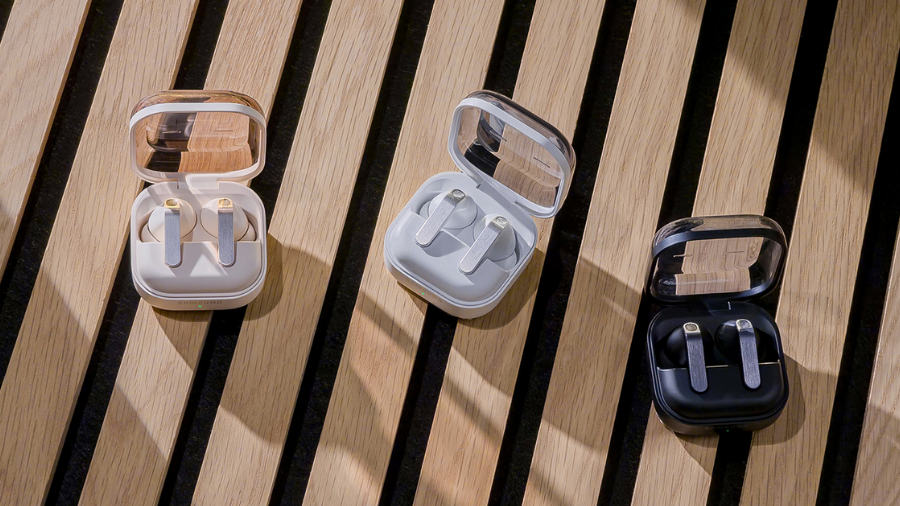 Tři otevřená nabíjecí pouzdra bezdrátových sluchátek Samsung Galaxy Buds4 v béžové, světle šedé a černé variantě položená na dřevěné lavičce, nasvícená přirozeným světlem.