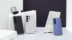 Zadní strana tří smartphonů řady Samsung Galaxy S26 v černé, světle šedé a modré barvě umístěných na kamenných blocích; vedle nich leží otevřené pouzdro sluchátek.