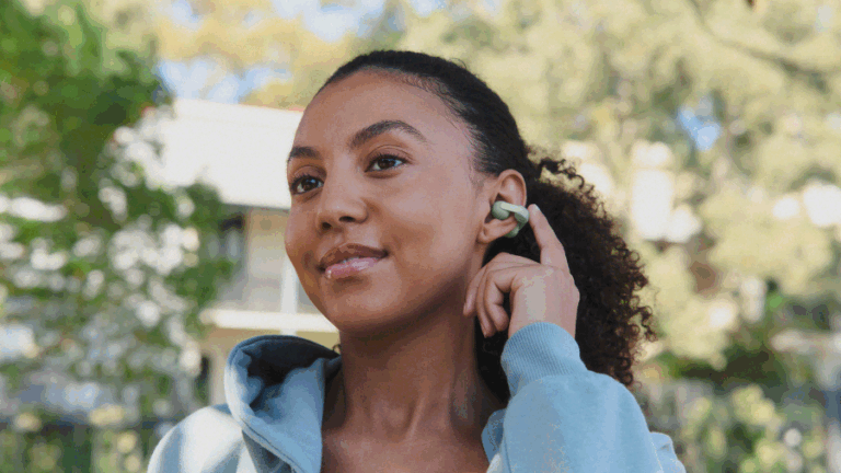 Portrét mladé usmívající se ženy s kudrnatými vlasy, která se dotýká nasazeného pastelově zeleného sluchátka Sony LinkBuds Clip. Žena má na sobě světle modrou mikinu s kapucí a nachází se venku, v pozadí je rozostřená zeleň a budova. Žena se dívá směrem doleva.