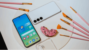 Telefon Samsung Galaxy S25 FE v bílé a černé barvy z přední i zadní strany včetně průhledného krytu. Vyfoceno vedle štětců a koblihy v růžových odstínech