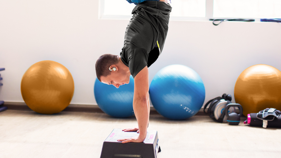 Sportovec provádějící stojku na rukou na step můstku se sluchátky Soundcore Sport X20. Snímek ilustruje extrémní stabilitu ušních háčků, které udrží sluchátka na místě i v obrácené poloze.