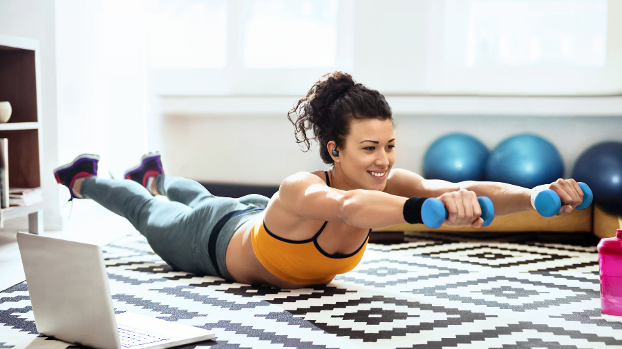 Žena cvičící doma na podložce s činkami, používající sluchátka Soundcore Sport X20 pro motivaci při sledování online tréninku na notebooku.