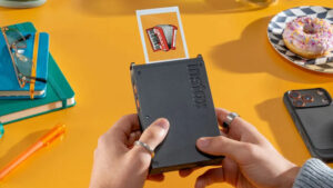 Detailní pohled na černou minitiskárnu Instax Mini Link+ v rukou uživatele, ze které právě vychází hotová fotografie s ilustrací akordeonu na pozadí žlutého stolu s doplňky.