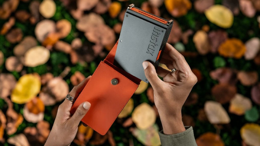 Pohled z ptačí perspektivy na ruce uživatele, který vkládá tmavě šedou minitiskárnu instax mini Link+ do stylového oranžového flipového pouzdra na podzimním pozadí s barevným listím.