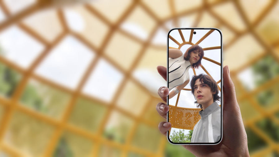 Detailní pohled na ruku držící smartphone POCO F8 Ultra, na jehož rozměrném displeji s minimálními rámečky je zobrazena ostrá a přirozená selfie dvou mladých lidí v moderním prostředí se žlutou geometrickou konstrukcí.