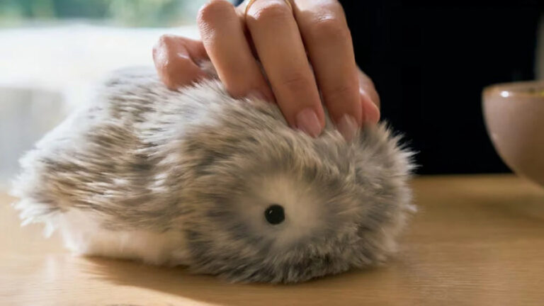 Detailní pohled na robotické zvířátko Moflin s šedou srstí, které na dřevěném stole hladí lidská ruka.