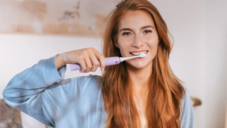 Na obrázku se usmívá mladá žena, která si v koupelně čistí zuby modelem TrueLife SonicBrush Clean70 UV Lavender. Tento snímek doplňuje naše srovnání sonických kartáčků a ukazuje elegantní fialový design v reálném použití.