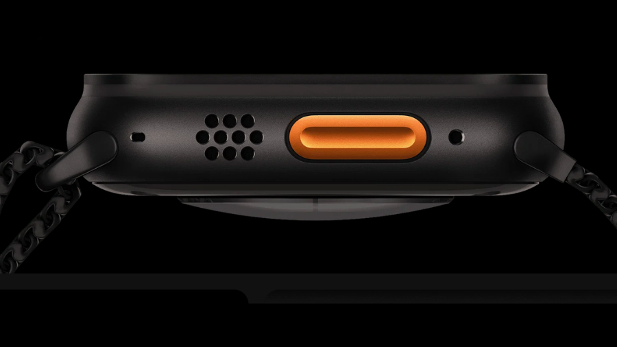 Detailní pohled z profilu na boční hranu černých hodinek Apple Watch Ultra 3 na tmavém pozadí. Uprostřed vyniká podlouhlé, jasně oranžové akční tlačítko, vedle kterého se nachází mřížka reproduktoru a otvory pro mikrofony. K hodinkám je připojen černý kovový řemínek.
