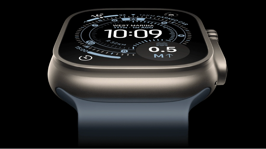 Detailní produktový pohled na displej a vrchní část hodinek Apple Watch Ultra 3 z nízkého úhlu na černém pozadí. Displej je aktivní, zobrazuje ciferník s kompasem, časem 10:09 a údaji o hloubce vody. Hodinky mají matné titanové pouzdro a šedomodrý řemínek.
