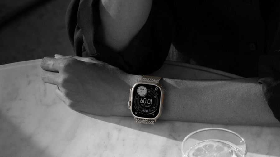 Černobílá lifestylová fotografie detailu ruky opřené o mramorový stůl, na jejímž zápěstí jsou hodinky Apple Watch Ultra 3 s kovovým řemínkem typu milánský tah. Vedle ruky stojí sklenice s vodou a citronem.