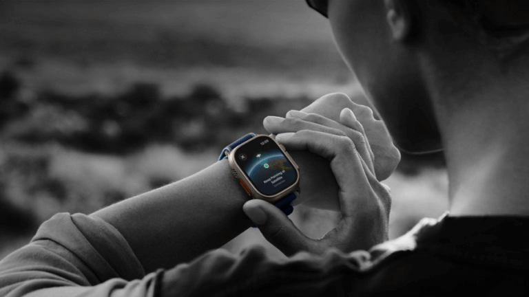 Černobílá fotografie z pohledu přes rameno ukazuje osobu, která se dívá na své zápěstí a ovládá hodinky Apple Watch Ultra 3. Hodinky mají titanové pouzdro, tmavě modrý řemínek a na jejich barevném displeji je zobrazena grafika aplikace pro připojení k satelitu s nápisem.