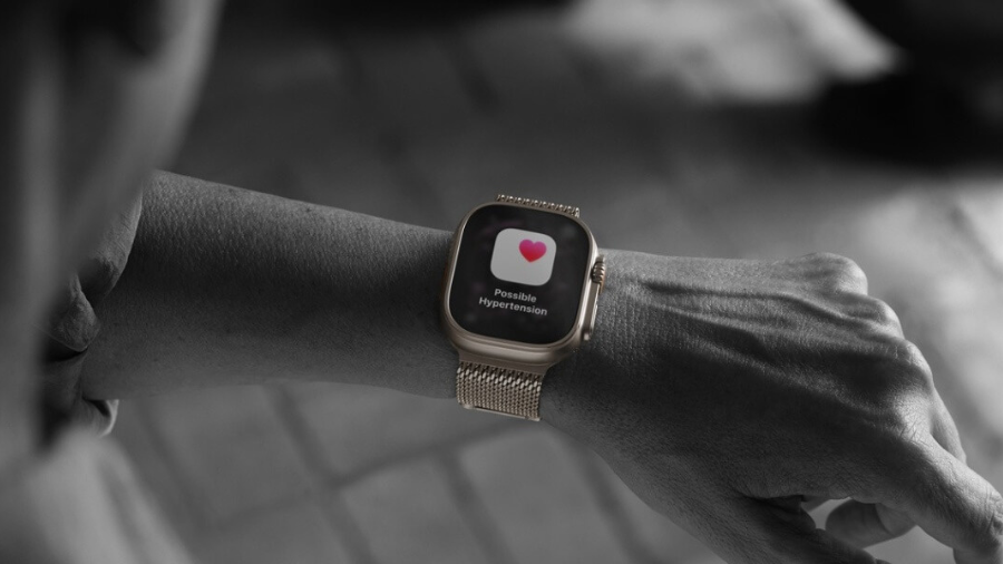 Černobílá lifestylová fotografie zápěstí s hodinkami Apple Watch Ultra 3 na kovovém řemínku milánský tah. Na jasném barevném displeji svítí zdravotní varování s ikonou srdce a textem „Possible Hypertension“, který upozorňuje na možný vysoký krevní tlak.