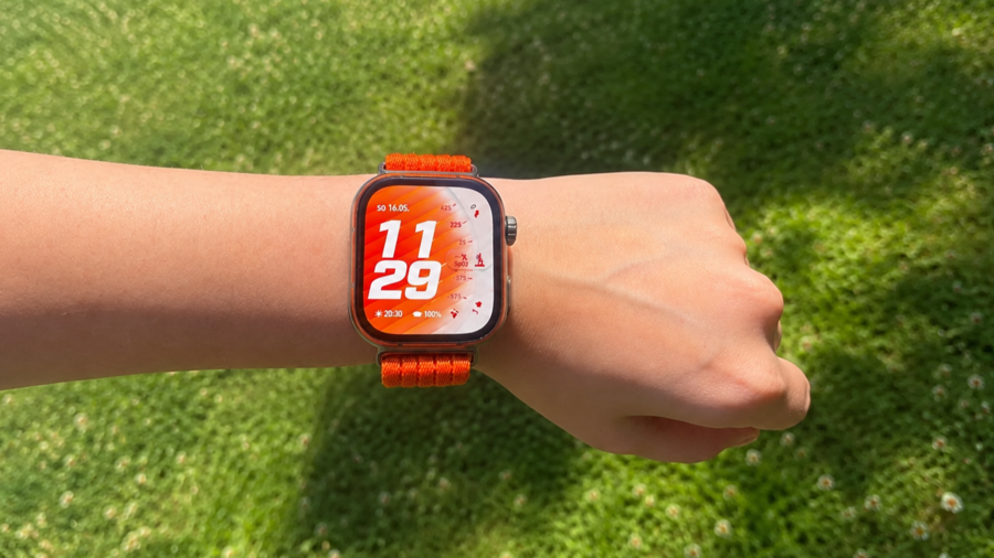 Detail chytrých hodinek Huawei Watch Fit 5 Pro s oranžovým řemínkem na zápěstí, zobrazujících ciferník s časem 11:29 na pozadí zeleného trávníku.