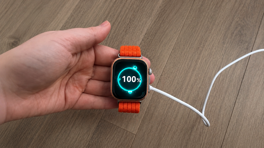 Ruka držící hodinky Huawei Watch Fit 5 Pro na nabíječce. Na displeji svítí tyrkysová ikona nabíjení se stavem 100 %. Hodinky mají výrazný oranžový řemínek.
