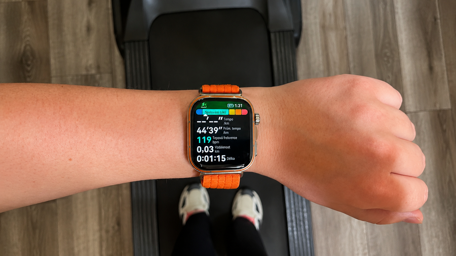 Pohled z perspektivy první osoby na chytré hodinky Apple Watch Ultra s oranžovým řemínkem na zápěstí. Na displeji v češtině běží fitness aplikace monitorující chůzi na běžeckém trenažéru, zobrazující tepovou frekvenci 119 bpm a zónu spalování tuků.