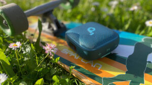 Modrý přenosný reproduktor soundcore Boom Go 3i položený na barevném longboardu v zelené trávě s sedmikráskami.