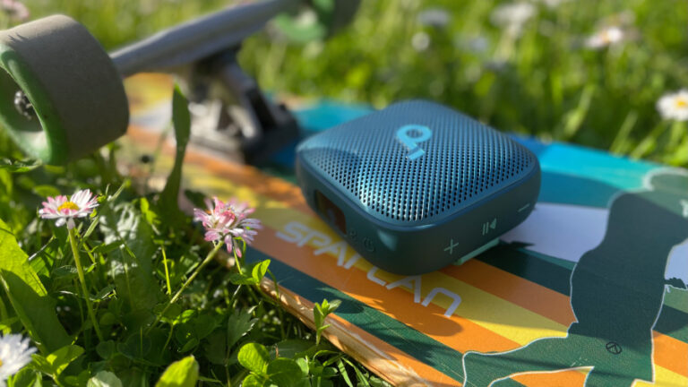 Modrý přenosný reproduktor soundcore Boom Go 3i položený na barevném longboardu v zelené trávě s sedmikráskami.