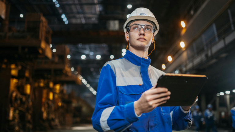 Mladý technik v ochranné helmě a pracovním oděvu sleduje data na tabletu v moderním průmyslovém závodě, což symbolizuje přerod obsluhy stroje v technologa.