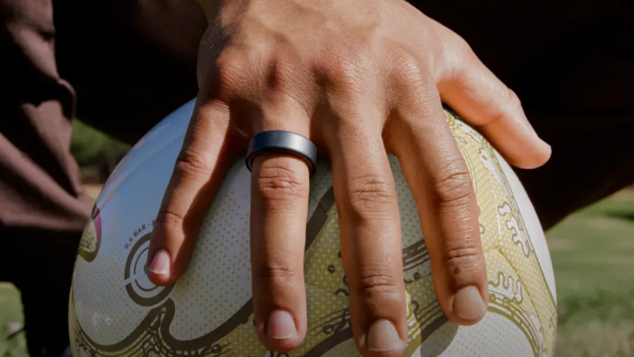 Detailní záběr na mužskou ruku s elegantním černým chytrým prstenem Oura Ring 5 položenou na fotbalovém míči jako originální dárek pro Den otců.
